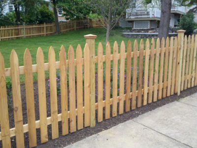 A simple wooden fence surrounding a lush green yard and bordering on a sidewalk